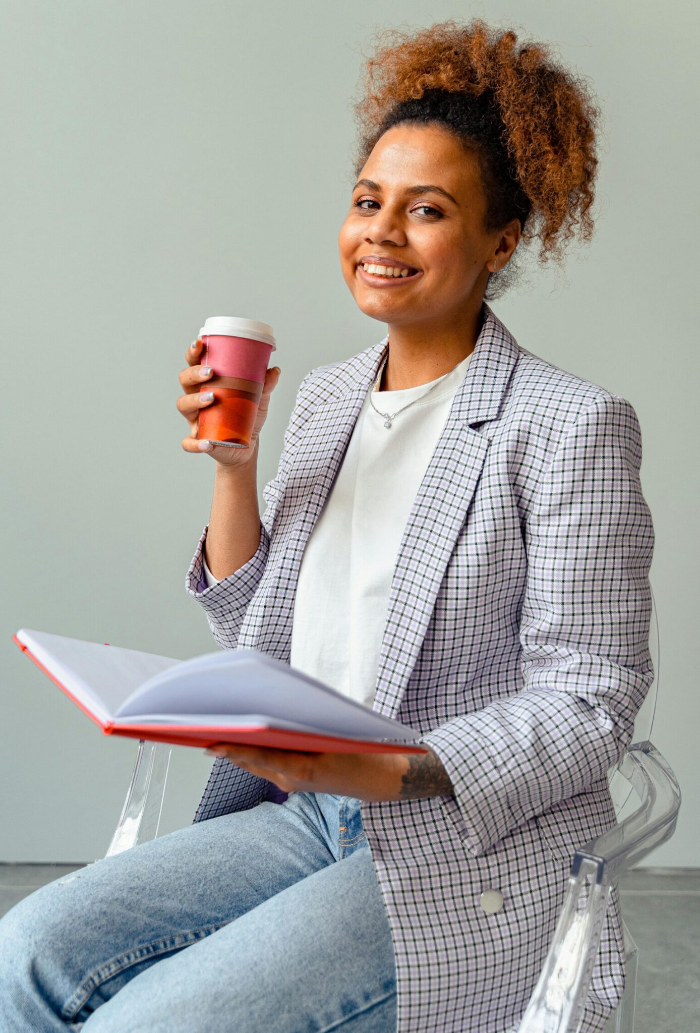 Smiling woman in blazer holds coffee cup and book, exuding confidence and warmth.